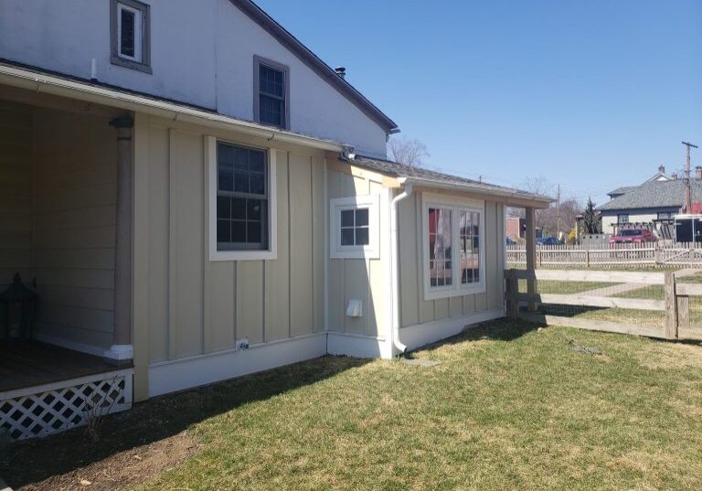 Exterior of a home featuring beige board and batten siding, white trim, and a small porch area under a clear blue sky.