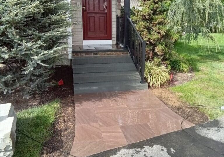 A small paver walkway with reddish-brown stones leads from an asphalt driveway to a set of dark grey steps at a home's entryway.