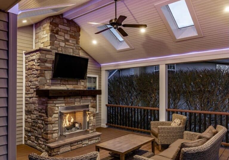 Covered deck with stone outdoor fireplace, mounted TV, wicker seating, fan, and purple accent lighting.