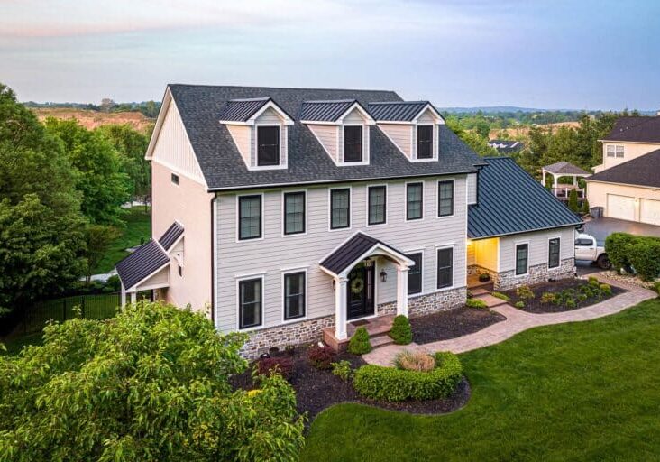 A large, modern Colonial-style home with light gray residential siding, stone veneer, black trim, and a dark metal roof.